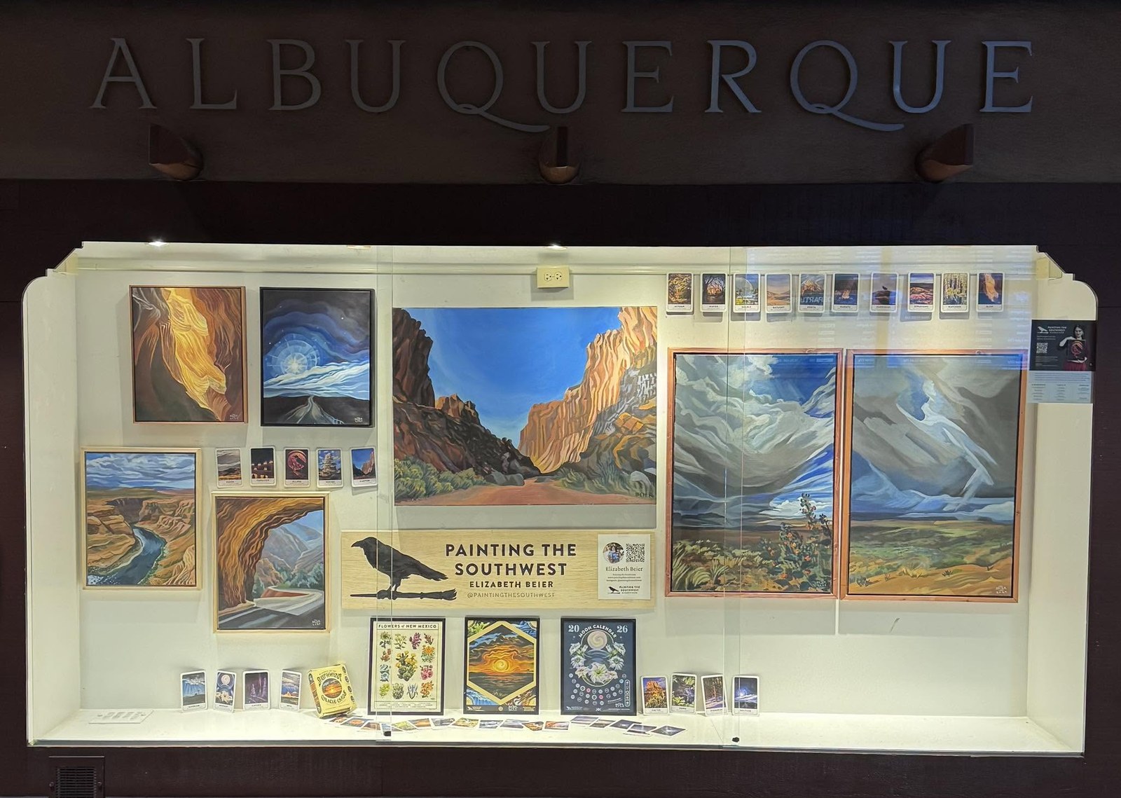 A lit display case beneath the ALBUQUERQUE letters at the airport, holding roughly a dozen framed Southwest landscape paintings, the Painting the Southwest raven sign with a QR code and artist card, a spread oracle deck, the Flowers of New Mexico print, a 2026 moon calendar and rows of postcards.